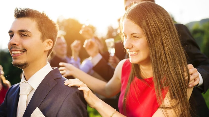 Woman in a red dress holds onto a suited man's shoulders in what appears to be a wedding celebration.