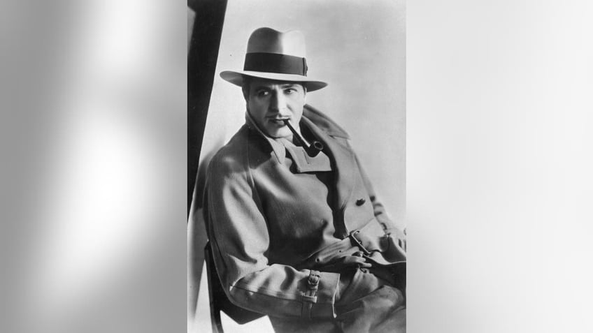 Warner Baxter wearing a trilby hat and coat, smoking a pipe.