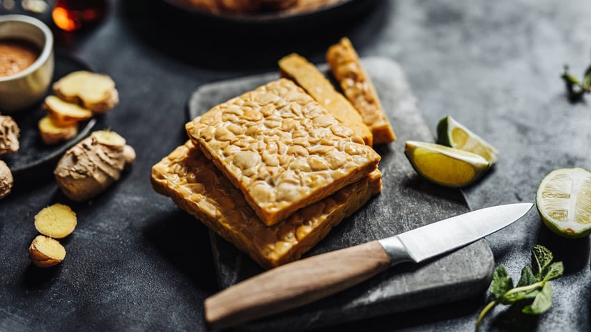 Close-up of tempeh on a cutting board with a kitchen knife and other ingredients on a table.