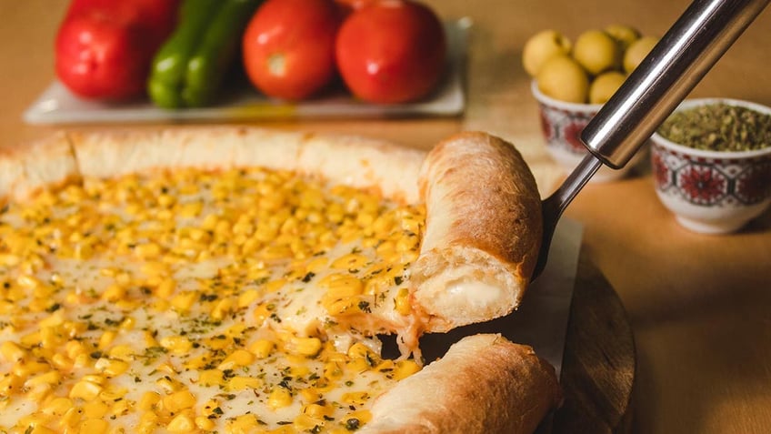 A slice of corn pizza is lifted up as it sits on a wooden board