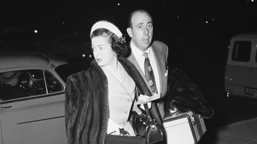 A close-up of Joan Bennett walking in a fur coat and suit next to a man in a suit.