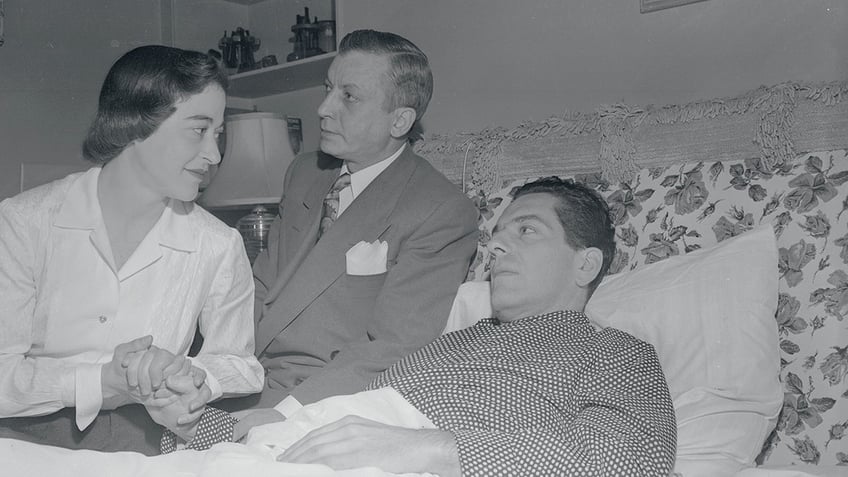 Jennings Lang laying in bed being consoled by a nurse as a man in a suit looks on.