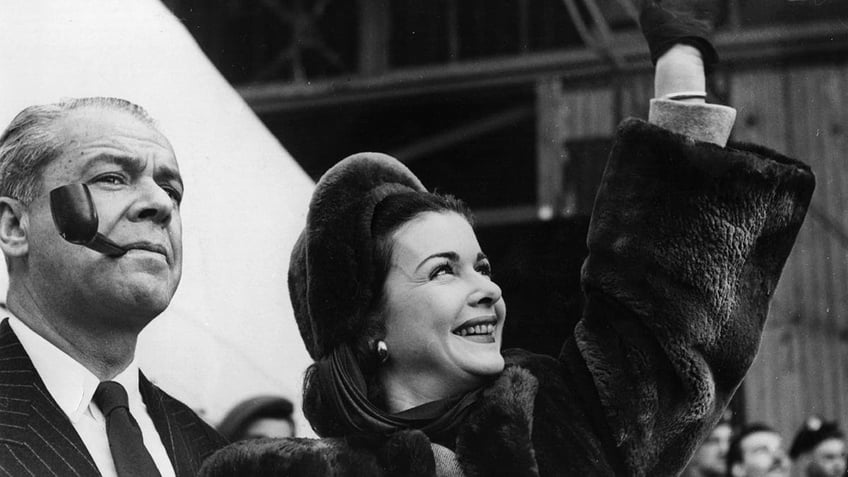 Walter Wanger smoking a pipe next to his wife Joan Bennett waving.