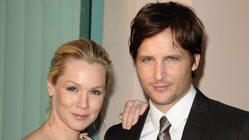 Jennie Garth puts her hand on husband Peter Facinelli's shoulder as they both soft smile/smirk on the carpet