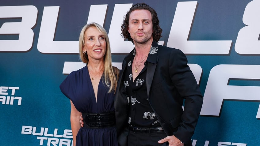 Sam Taylor-Johnson in a navy dress smiles on the carpet with husband Aaron Taylor-Johnson in a black printed shirt and black suit jacket