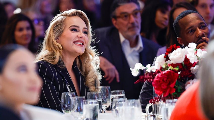 Adele in a pinstripe jacket sits at a table beaming next to boyfriend Rich Paul