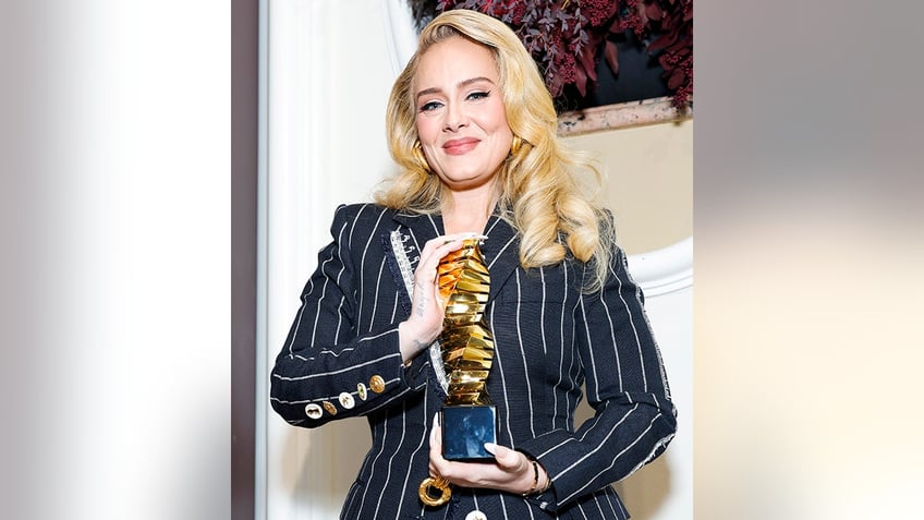 Adele soft smiles while she holds up her award from The Hollywood Reporter in a blue pinstripe jacket