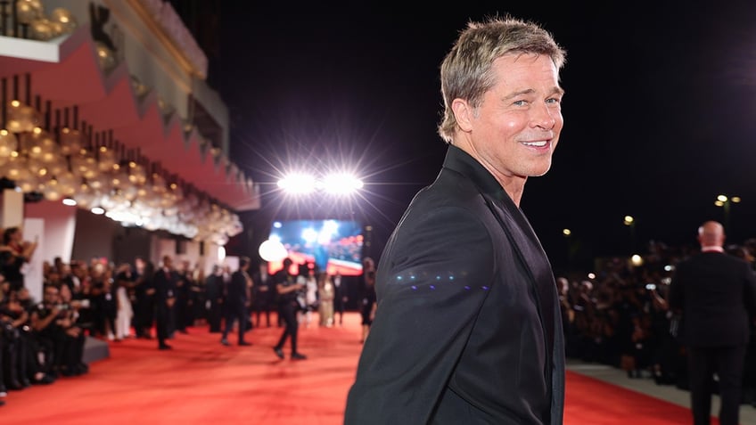 Brad Pitt in a black suit walks on the red carpet and looks towards the camera in Venice