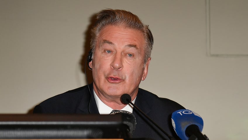 Alec Baldwin in a black suit speaks into a microphone during a press conference at the Torino Film Festival