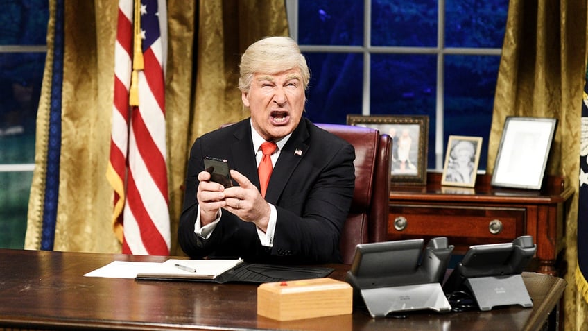 Alec Baldwin dressed as President Donald Trump in a black suit and red tie holds a phone in his hands and looks upset