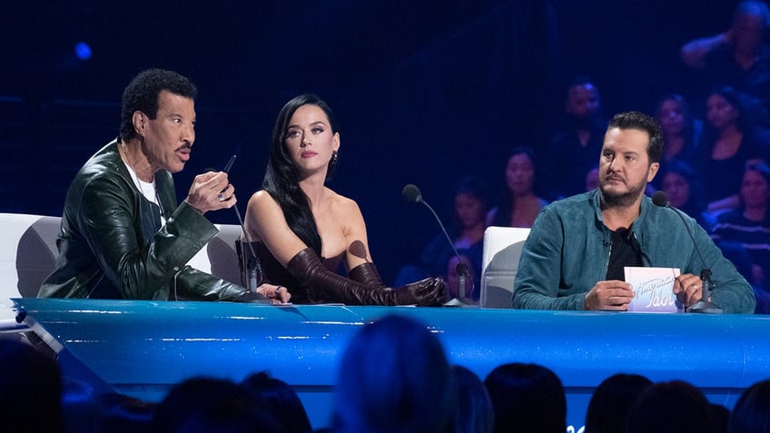 Lionel Richie speaks with a pen seated behind the judge's desk on "American Idol" with Katy Perry next to him and Luke Byran looking over at him