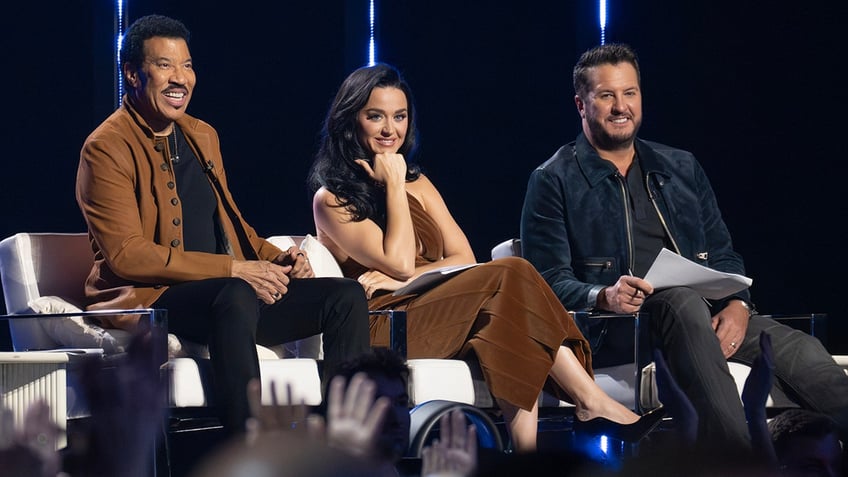 Lionel Richie in a brown jacket sits in a chair next to Katy Perry in brown pants next to Luke Bryan for 