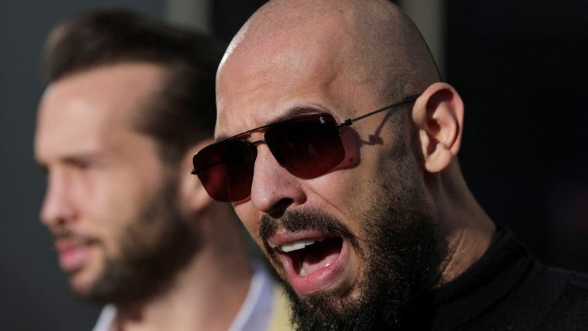 Andrew Tate speaking in a closeup image, he is wearing a black shirt and sunglasses, is bald and has a bushy goatee