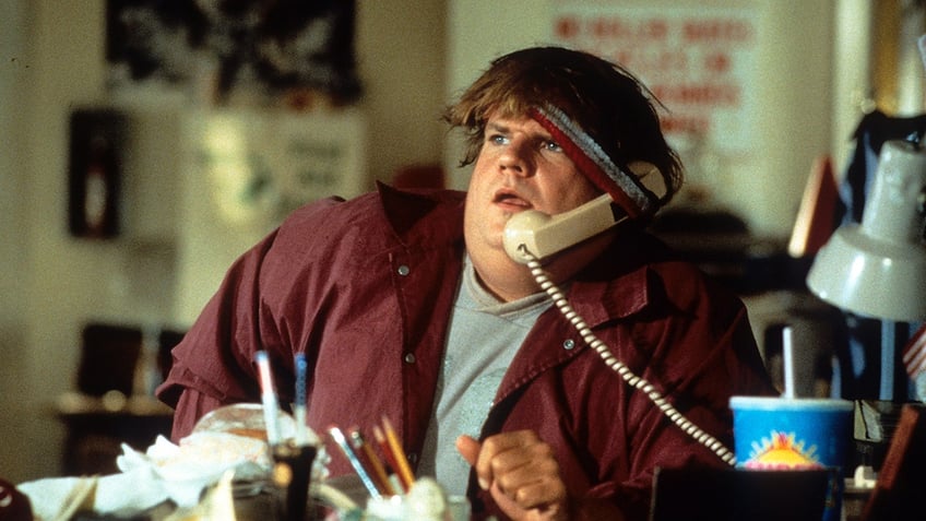 Chris Farley holding a phone receiver underneath his headband to keep it propped up to his ear