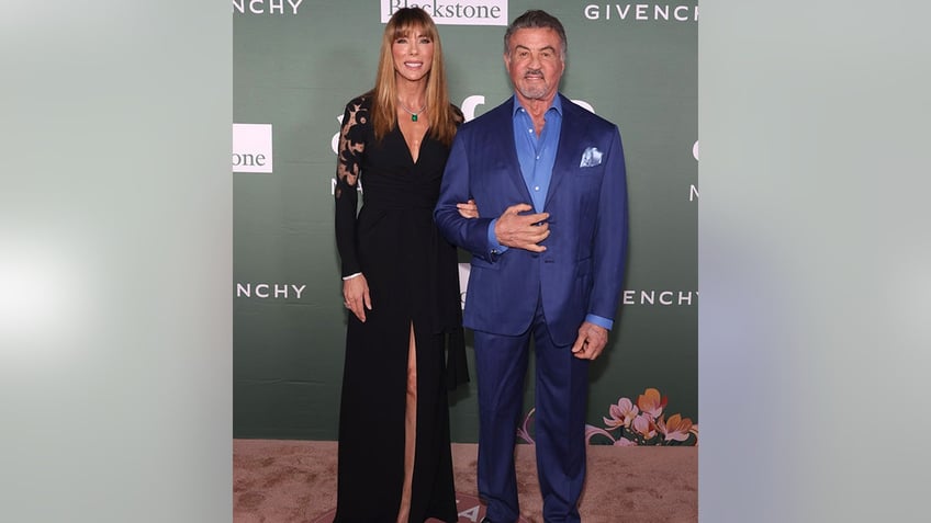 Jennifer Flavin in a black dress with a slit down the middle poses with husband Sylvester Stallone in a bright royal blue jacket
