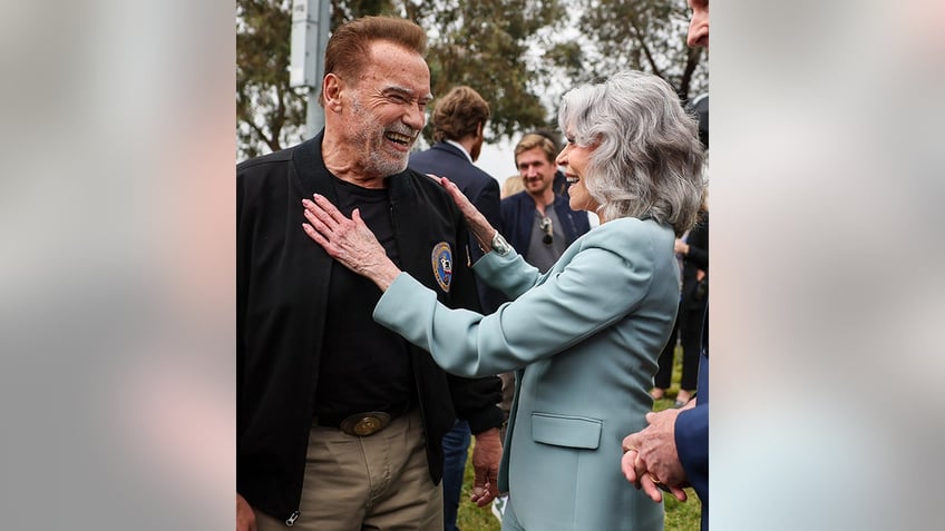 Arnold Schwarzengger smiles in his black suit and tan khaki pants as he is greeted by Jane Fonda in a blue suit