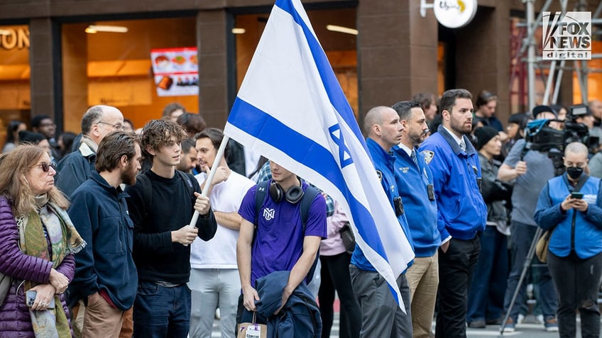 crowd on sidewalk, man holds Israeli flag in left-center of photo