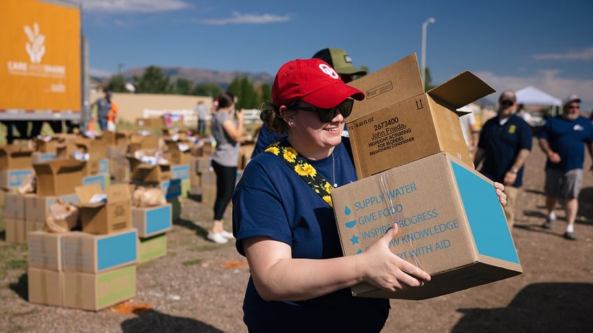 Woman in blue shirt and red hat walking with boxes of donated food.