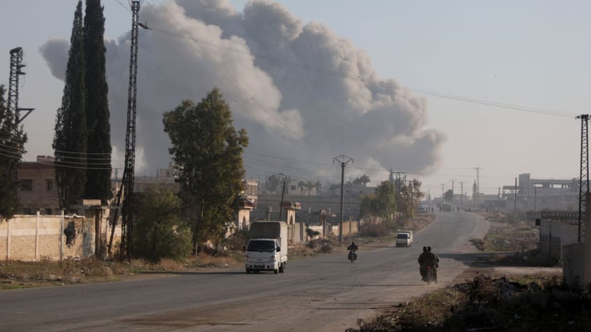 Fighters enter the Rashidin district on the outskirts of Aleppo on their motorbikes with smoke billowing in the background during fighting on November 29, 2024, as Hayat Tahrir al-Sham (HTS) jihadists and allied factions continue their offensive in the Aleppo province against government forces. 