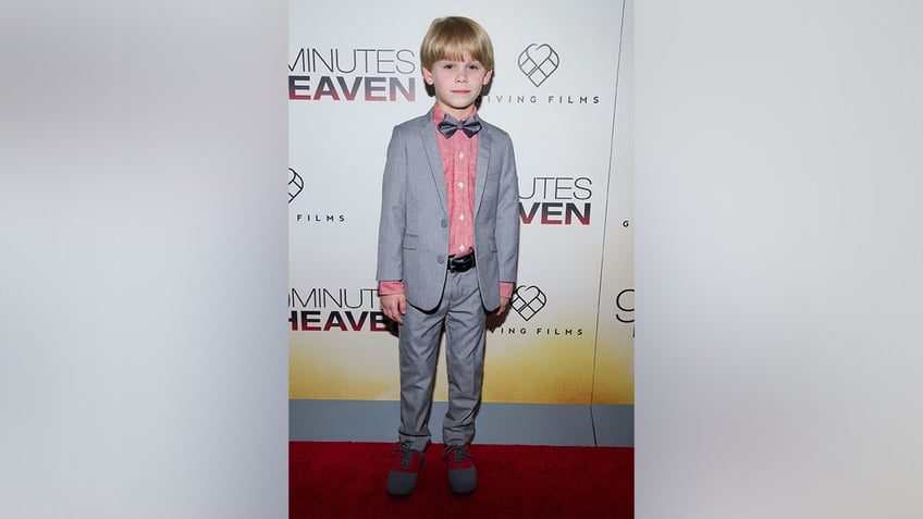 Hudson Meek in a grey two piece suit and red button down looks stoic as a child on the carpet