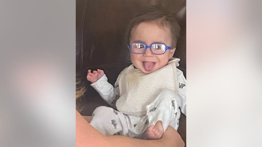 Baby Liam smiles and sticks out his tongue while wearing his new glasses.