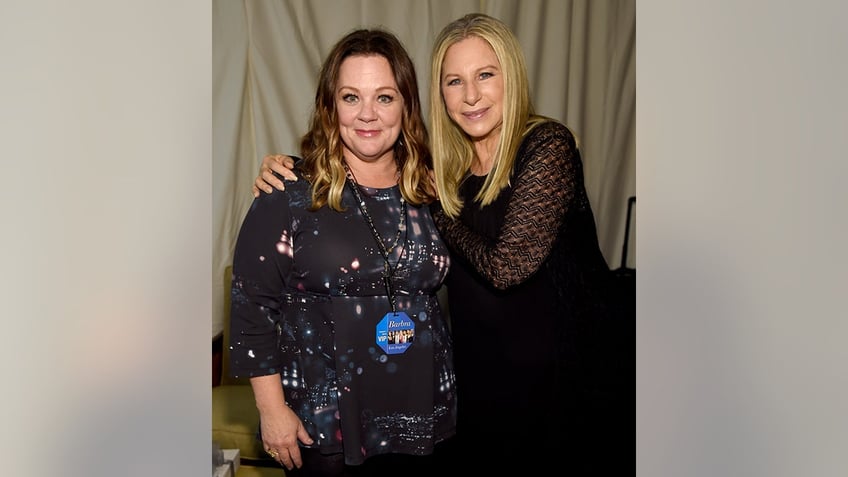 Melissa McCarthy in a black patterned dress has her shoulders hugged by Barbra Streisand in black