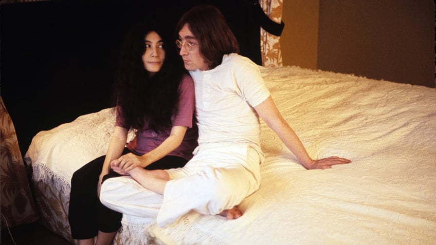 Yoko Ono looking at John Lennon who is wearing all white while they sit on a bed.