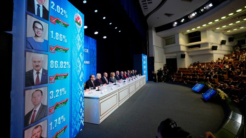 Results from Belarus' 2024 election are shown on screens flanking a long table at which Belarusian bureaucrats sit.