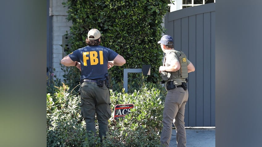 A man in a navy blue FBI jacket stands with his back to the camera next to another gentleman in a green FBI jacket outside Ben Affleck's home