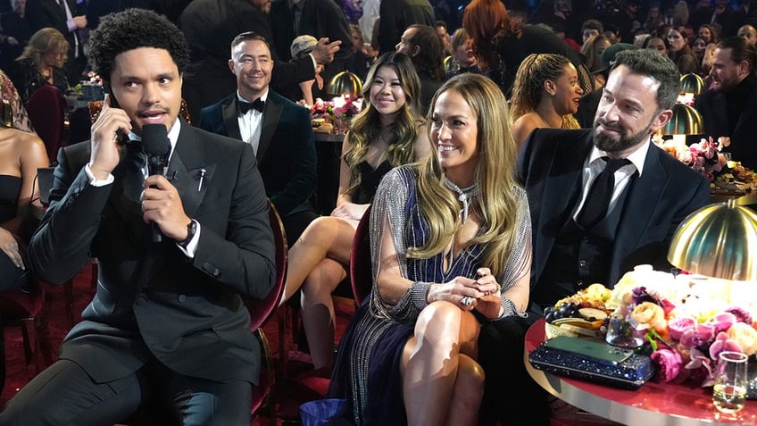 Trevor Noah speaks into the microphone while hosting the Grammys as Jennifer Lopez, smiling, and Ben Affleck look on from the table next to him