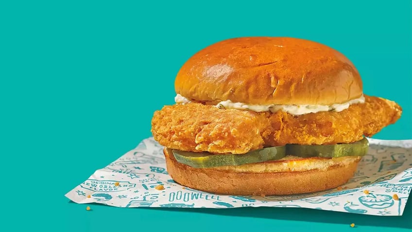 A Popeyes Cajun Flounder Fish Sandwich is on display in front of a teal background.