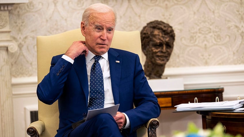 A seated President Biden tugs at his collar
