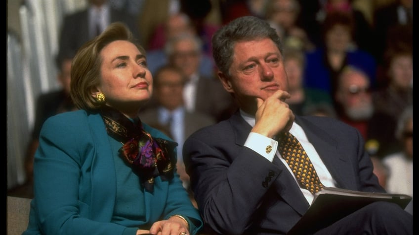 Pres. Bill & Hillary Rodham Clinton sitting together, listening to spkr., framed by audience attending health care event.