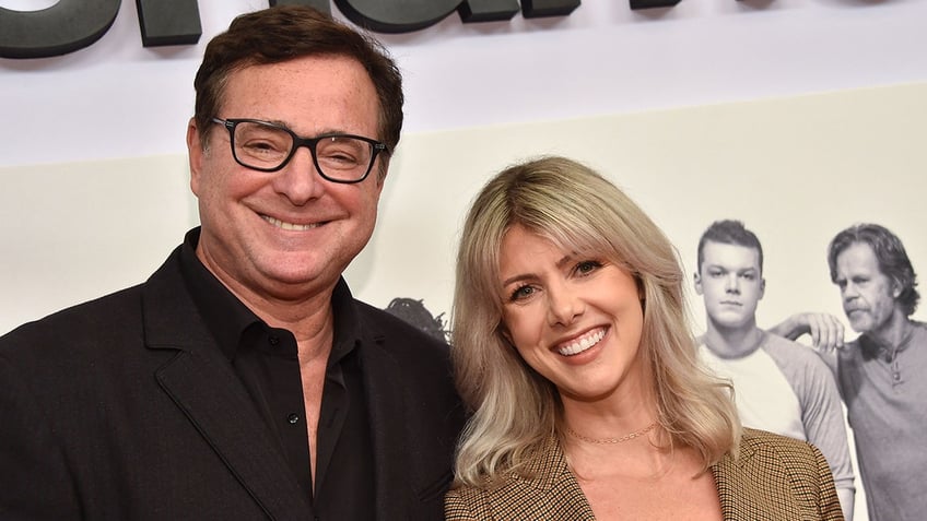 Bob Saget in a black shirt with black frames smiles next to wife Kelly Rizzo in a tan jacket on the red carpet