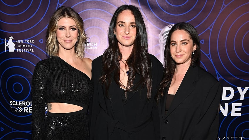 Kelly Rizzo in a black cut out dress soft smiles on the carpet with Bob Saget's daughters Lara Saget and Aubrey Saget, both of whom are also wearing black