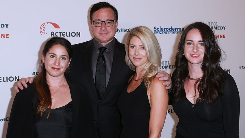 Bob Saget in a black suit holds on to his daughter and wife Kelly Rizzo, as another daughter stands next to Rizzo, all wearing black