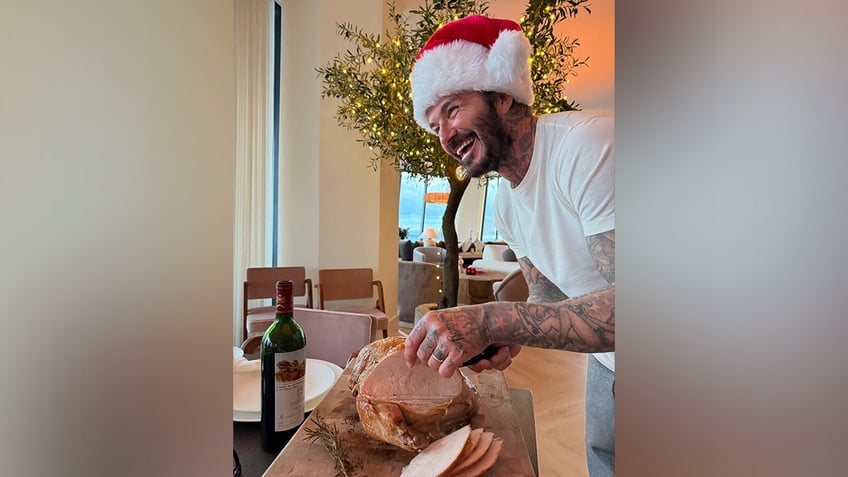 David Beckham wearing a Santa hat smiles in a white t-shirt, grey sweatpants, and a santa hat while carving turkey