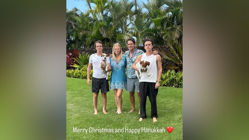 Matthew Lowe holds a dog in a white t-shirt and dark camo shorts next to mother Sheryl in a blue tunic dress next to Rob in a tropical patterned shirt and shorts next to Johnny holding a dog wearing a white tank top and black pants