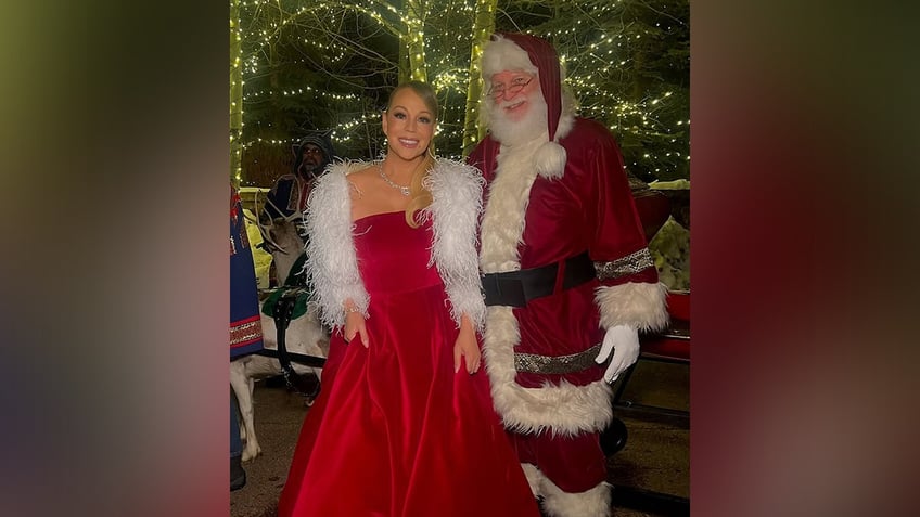 Mariah Carey in a velvet red strapless dress and white furry cardigan smiles next to Santa Claus