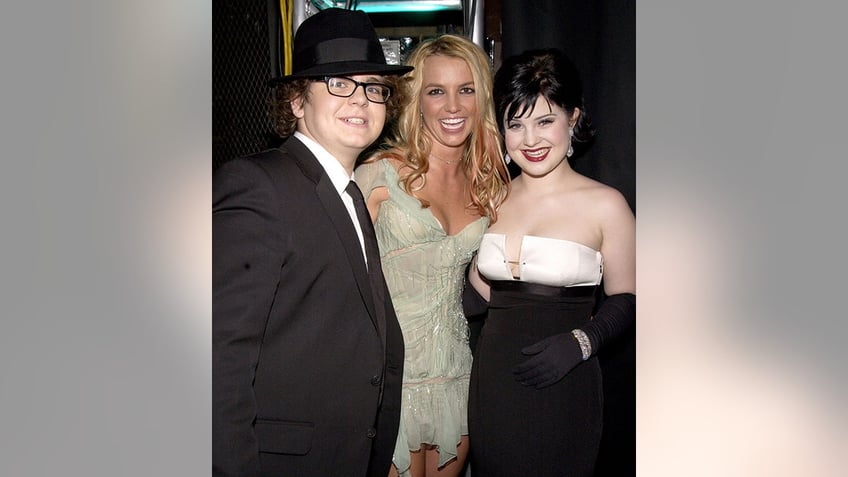 Jack Osbourne in a black suit and top hat smiles with Britney Spears in a light green dress and Kelly Osbourne in a white and black dress