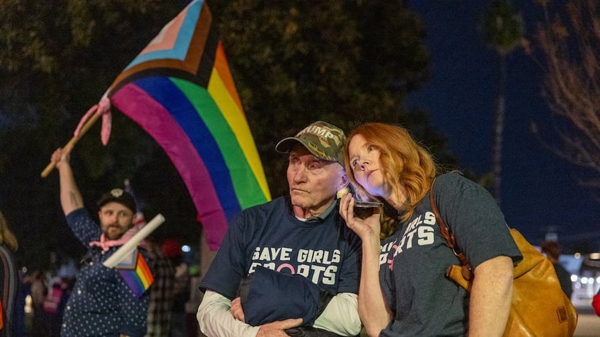 Transgender athlete supporter Kyle Harp, left, of Riverside holds the progress  pride flag as "Save Girls Sports" supporters Lori Lopez and her dad Pete Pickering, both of Riverside, listen to the debate as they join the overflow crowd converging outside the Riverside Unified School District meeting Thursday night to debate the rights of transgender athletes to compete in high school sports Thursday, Dec. 19, 2024. 