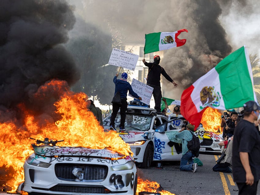 Demonstrators wave flags from atop a wrecked car, standing beside burning Waymo vehicles,