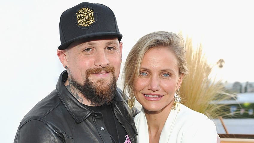 Cameron Diaz in a white blazer smiles for a photo with husband Benji Madden in a black leather jacket and black hat