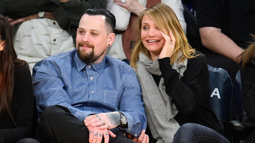 Benji Madden in a denim shirt puts his hands on his knees while sitting courtside at a basketball game with Cameron Diaz
