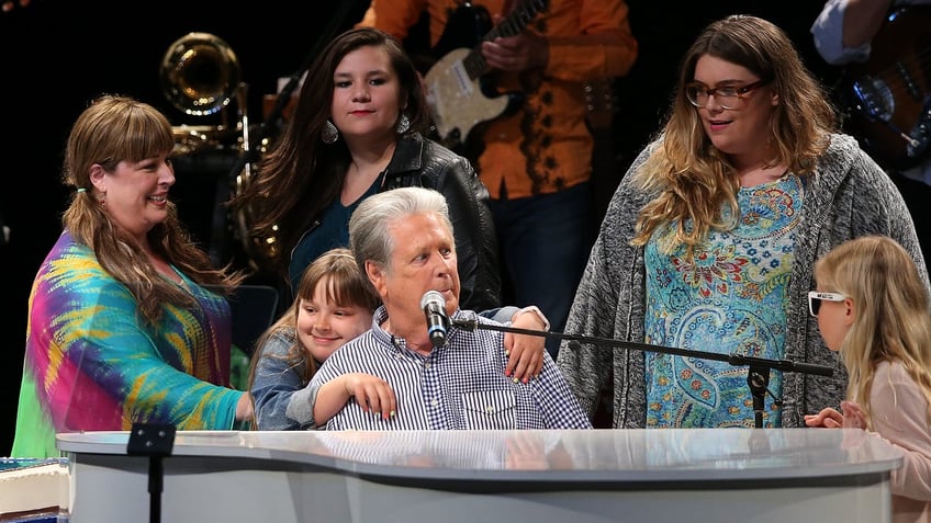 lola hugging her grandfather brian wilson as he plays the piano next to with mother carnie at brians birthday party