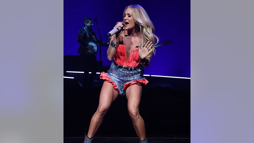 Carrie Underwood in a red bustier top and jean shorts with red trim sings passionately into the microphone
