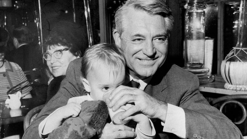 Cary Grant smiling and holding onto his daughter Jennifer as she drinks from a cup