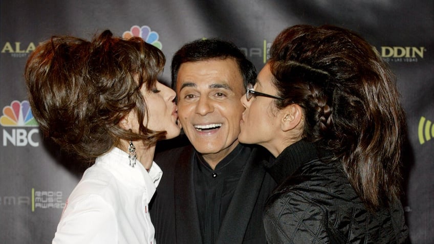 Casey Kasem smiling as his two daughters kiss him on each cheek.