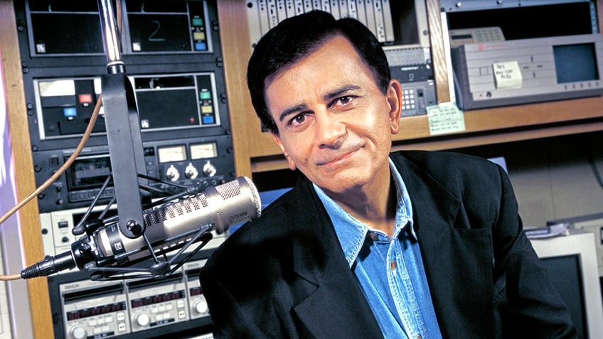 Casey Kasem posing next to a mic in a dark blazer and a denim shirt.