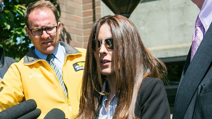 Kerri Kasem wearing glasses speaking in front of a row of mics